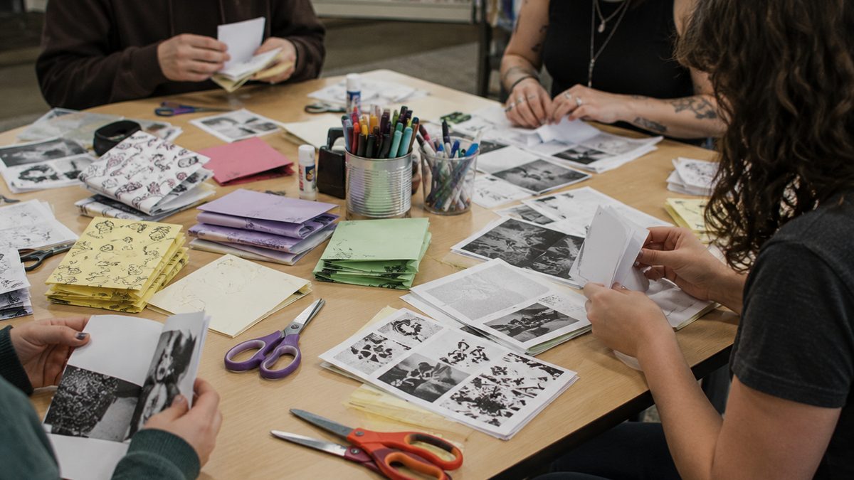 Hands assemble folded paper zines on a library workshop table with markers and scissors.
