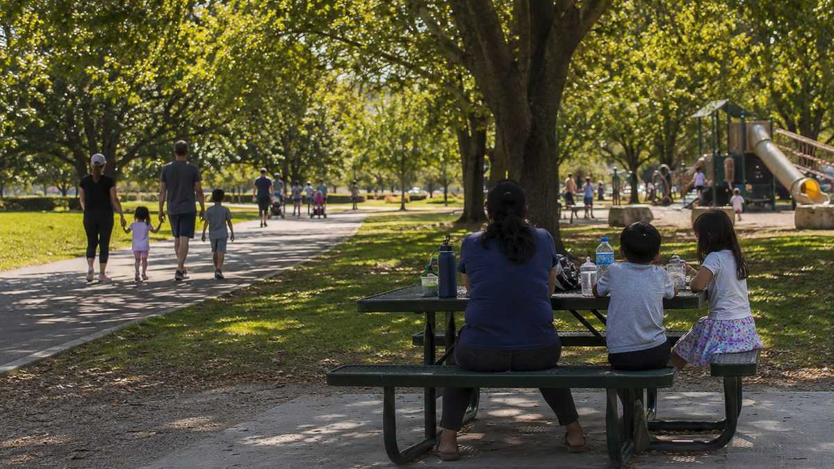 Families use a public park with picnic tables, shade trees, a playground, and walking paths.
