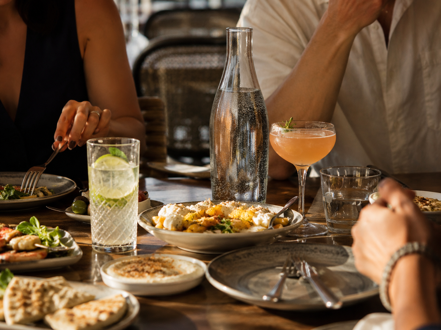 A restaurant table with a cocktail, a nonalcoholic drink, water, and shared appetizers in a calm evening setting.