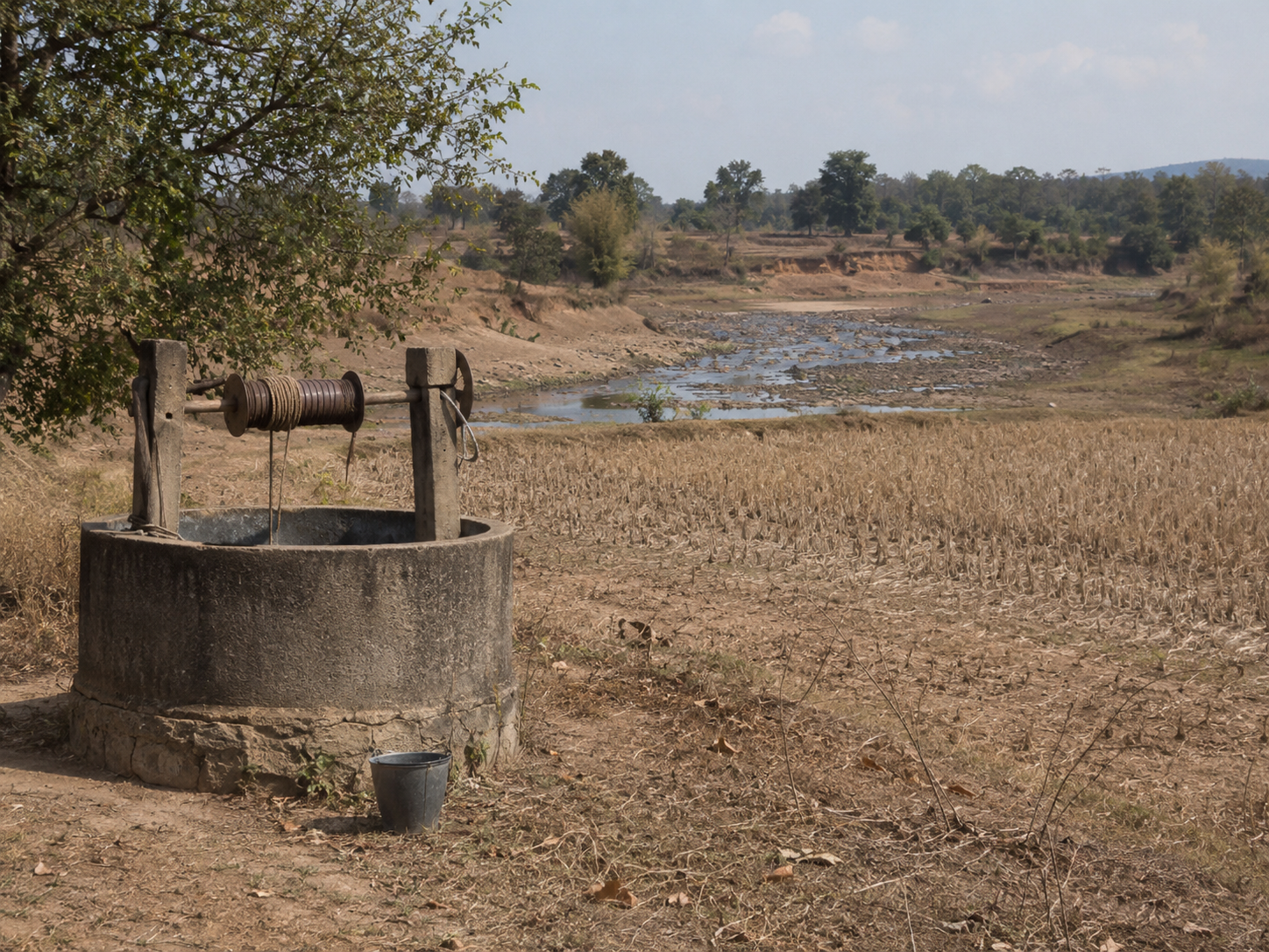 A rural water well stands near a dry field and shallow streambed.