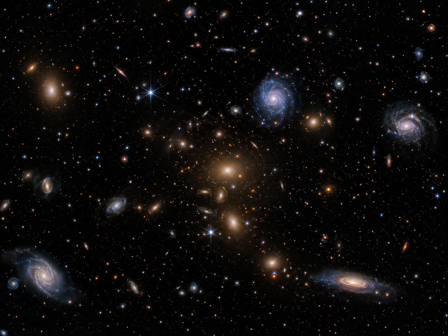 A galaxy cluster with spiral and elliptical galaxies against a dark background.