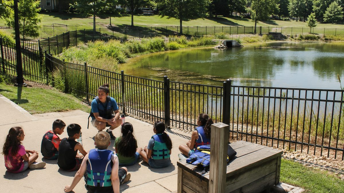 Children learn water safety near a fenced community pond with an instructor nearby.